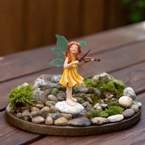 May include: A miniature figurine of a fairy with long red hair, a yellow dress, and green wings, playing a brown violin. The fairy stands on a white stone base surrounded by small gray pebbles and patches of green moss on a circular tray.