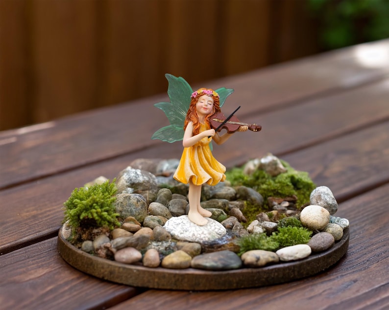 May include: A miniature figurine of a fairy with long red hair, a yellow dress, and green wings, playing a brown violin. The fairy stands on a white stone base surrounded by small gray pebbles and patches of green moss on a circular tray.