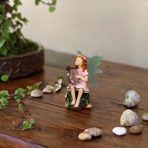 May include: A small figurine of a fairy with orange hair and translucent green wings, wearing a pink dress, sitting on a brown tree stump while playing a brown lyre decorated with small pink flowers. The fairy has eyes closed in a peaceful expression.