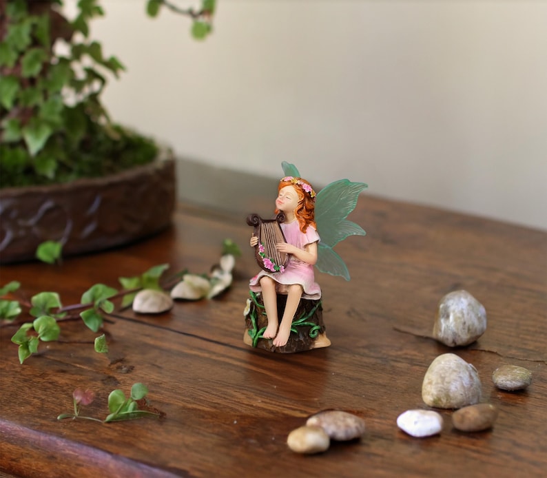 May include: A small figurine of a fairy with orange hair and translucent green wings, wearing a pink dress, sitting on a brown tree stump while playing a brown lyre decorated with small pink flowers. The fairy has eyes closed in a peaceful expression.
