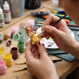 May include: A small figurine of a fairy with long orange hair, wearing a yellow dress and floral crown, holding a miniature violin. The fairy has translucent light green wings and is being painted with a fine-tipped brush and acrylic paints.