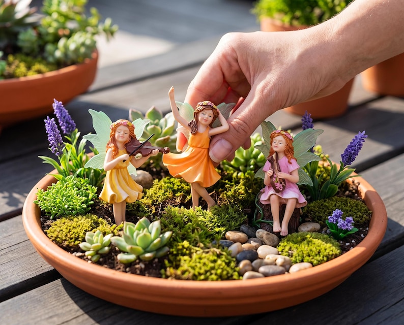 May include: Three miniature fairy figurines with wings, one in a yellow dress playing a violin, one in an orange dress dancing, and one in a pink dress playing a lute, arranged in a round terracotta planter with succulents, moss, and pebbles.