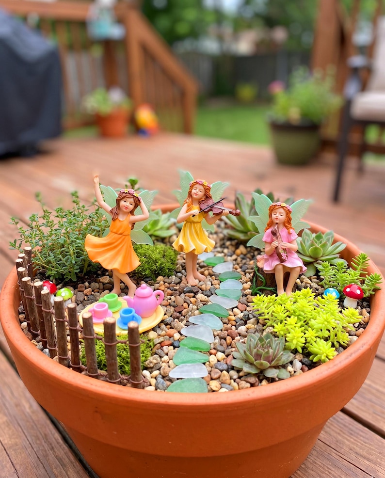 May include: A terracotta planter containing a miniature garden with three fairy figurines, a small wooden fence, a path of green glass stones, succulents, and a tea set. One fairy wears orange, one yellow, and one pink, with small mushrooms nearby.