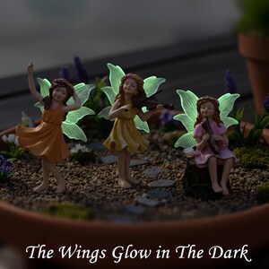 May include: Three fairy figurines with glowing wings in a terracotta bowl. One dances in an orange dress, one plays a violin in a yellow dress, and one sits on a stump playing a flute in a pink dress. Text reads: The Wings Glow in The Dark.