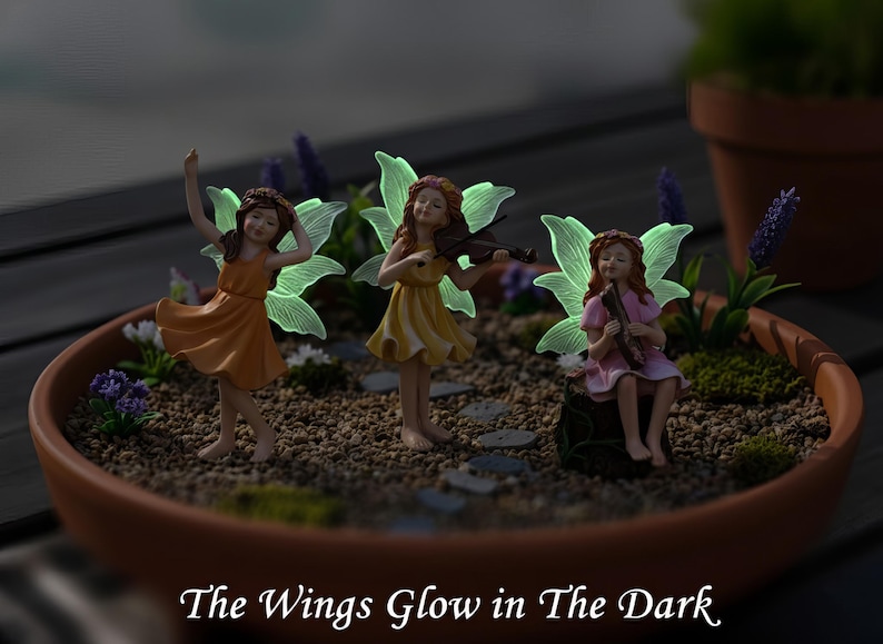 May include: Three fairy figurines with glowing wings in a terracotta bowl. One dances in an orange dress, one plays a violin in a yellow dress, and one sits on a stump playing a flute in a pink dress. Text reads: The Wings Glow in The Dark.