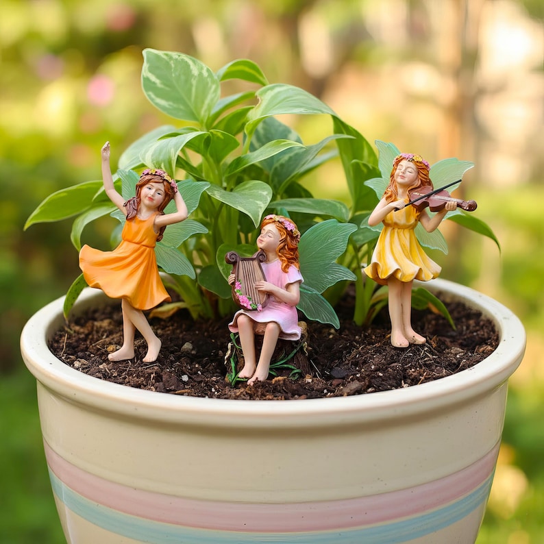 May include: Three small fairy figurines placed in a potted plant. One fairy wears an orange dress and dances, one sits on a stump playing a pink lyre in a pink dress, and one stands in a yellow dress playing a brown violin. Each wears a floral crown.