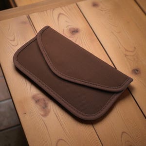 May include: A brown, rectangular case with a curved flap closure. The case is made of a textured fabric and features a stitched border. The case is resting on a wooden surface.