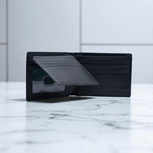 May include: Open black leather wallet with several card slots and a clear ID window. The wallet is shown open, displaying its interior design and storage capacity. The background is a marble surface.