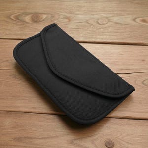 May include: A black, rectangular, fabric case with a curved flap closure. The case is made of a textured material and has a stitched border. The background is a light brown wooden surface.