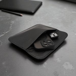 Op de afbeelding: Zwarte leren etui met een telefoon en autosleutel. De etui is open en toont de inhoud. Een zwarte telefoonhoes en een autosleutel zijn zichtbaar. Een zwarte laptop en een potlood staan op de achtergrond.
