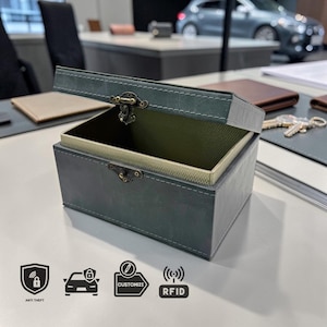 May include: A dark teal leather box with a hinged lid, open to show a lighter interior. The box has a decorative clasp and white stitching. Icons for anti-theft, car security, customization, and RFID are visible.