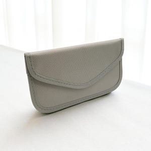 May include: A light gray, rectangular glasses case with a curved flap closure. The case is made of a textured fabric and features a stitched border. The design is simple and minimalist, suitable for protecting eyewear.