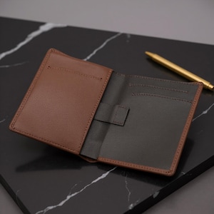 May include: Open brown leather wallet with a dark interior and a small tab closure. The wallet has a contrasting brown border and visible stitching. A gold pen rests on the surface next to the wallet.