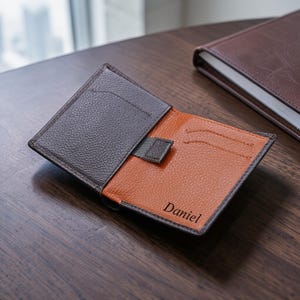 May include: Open brown and dark brown leather wallet with the name "Daniel" printed on the inside. The wallet has card slots and a dark green strap closure. A brown leather notebook is in the background.
