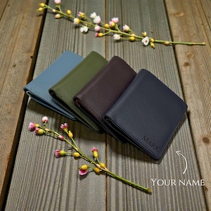 May include: Four leather wallets in blue, green, brown, and navy, arranged on a wooden surface. The navy wallet is embossed with the name "MARK". Delicate floral sprigs with pink and yellow blossoms add a touch of colour.