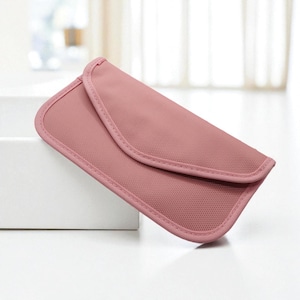 May include: A dusty rose-colored, rectangular pouch with a curved flap closure. The edges are neatly trimmed with a matching pink binding. The pouch is made of a textured fabric and appears to be empty. The pouch is resting on a white surface.
