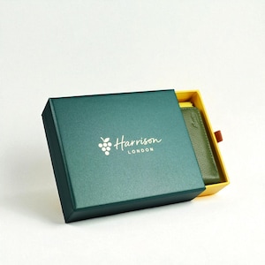 May include: A teal green gift box with the name "Harrison LONDON" in white script. The box is open, revealing a green leather wallet inside a yellow interior. The box has a white background.