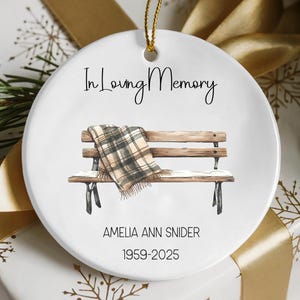 May include: A white ceramic ornament with a gold string for hanging. The ornament features a watercolor illustration of a park bench with a blanket and the text "In Loving Memory." Below the bench are the name and dates "AMELIA ANN SNIDER 1959-2025."