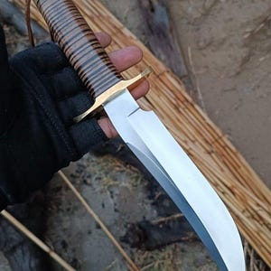 Puede incluir: Un cuchillo grande de acero pulido con una hoja curva. El mango está envuelto en cuero marrón y negro, con una guarda y pomo de latón. Una correa de cuero está sujeta al mango. El cuchillo se sostiene en una mano enguantada.