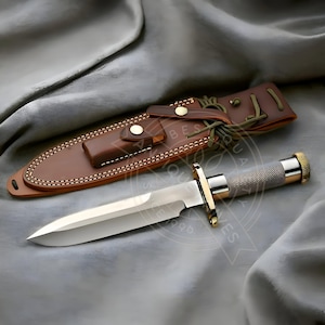May include: A polished steel knife with a sharp blade, gold accents, and a textured handle, presented with a brown leather sheath. The sheath has decorative stitching and a button closure. The knife and sheath are displayed on a gray fabric.