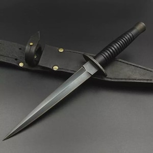 May include: A black dagger with a double-edged blade, a ridged handle, and a crossguard. The dagger is in a black leather sheath with brass rivets. The design suggests a tactical or combat knife.