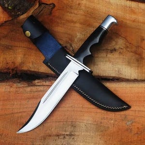 May include: A hunting knife with a polished silver blade and a black handle, partially sheathed in a black leather case with a blue interior. The knife is set against a wooden background.
