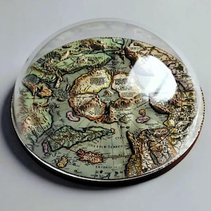 May include: A circular, three-dimensional relief map of the Arctic region encased under a clear, domed glass cover. The map features raised topographical details of landmasses and oceans in muted earth tones, with printed text labels throughout.