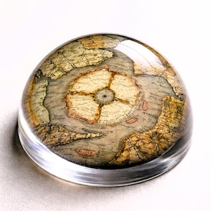 Puede incluir: Un pisapapeles transparente en forma de cúpula con un diseño de mapa antiguo. El mapa presenta tonos beige, marrón y gris, que representan continentes y detalles geográficos. El pisapapeles está hecho de vidrio transparente.