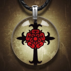 May include: A round pendant necklace featuring a black cross design with a red rose in the center. The pendant is encased in a clear, glass-like material and hangs from a black braided cord. The pendant has a silver-colored bail.