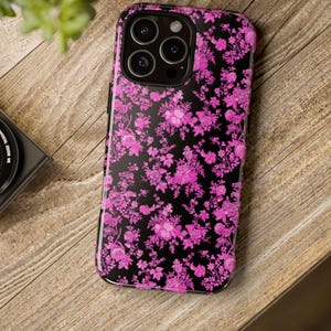 Peut inclure: Coque de smartphone noire ornée d'un motif floral rose vif. L'étui présente une finition brillante et est conçu pour protéger l'appareil. Le motif floral est un arrangement répété de fleurs et de feuilles.