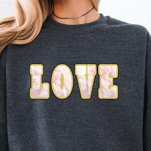 May include: Dark heather gray sweatshirt with the word "LOVE" in a gold outline with a pink and white floral design. The sweatshirt is a long-sleeved crew neck style.