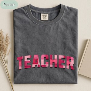 Peut inclure: Un t-shirt gris foncé avec le mot "TEACHER" en lettres patchwork roses et rouges sur le devant. Le t-shirt a un col rond et une étiquette "Comfort Colors".
