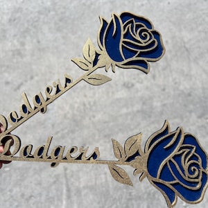 Peut inclure: Deux décorations en forme de rose en bois. Chacune présente une rose bleue avec des accents dorés et le mot "Dodgers" en écriture cursive. Les décorations sont tenues sur un fond gris.