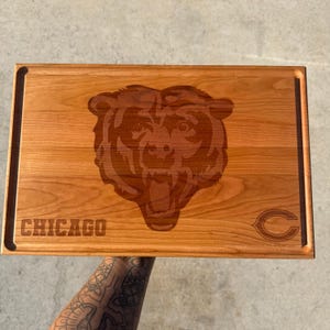 May include: A rectangular wooden cutting board with a natural light brown grain. The center features a laser-engraved bear head graphic. The word CHICAGO is engraved on the bottom left, and a C logo is engraved on the bottom right corner.