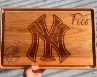 May include: A rectangular wooden cutting board with a carved New York Yankees logo, the name "Fico", and a Yankees logo. The board is light brown with a darker brown border and engraved details.