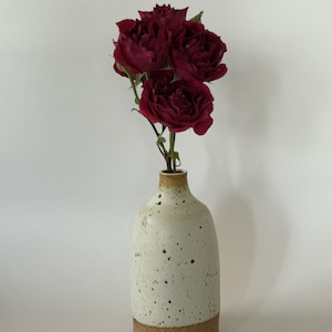 May include: A ceramic vase with a speckled white finish and a cork base holds a small bouquet of deep red roses. The vase has a rounded shape and a narrow neck, perfect for displaying flowers. The roses are in full bloom.