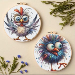 May include: Two whimsical cross-stitch embroidery hoops featuring cartoon birds. One bird has gray and blue feathers, the other has blue, purple, and orange feathers. Both have large, expressive eyes and are set in wooden hoops.