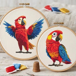 Puede incluir: Dos diseños de loros de punto de cruz en aros de madera. Un loro tiene las alas azules y amarillas extendidas, el otro está de pie. Ambos loros son rojos, amarillos y azules. Hilos de bordar y un dedal en primer plano.