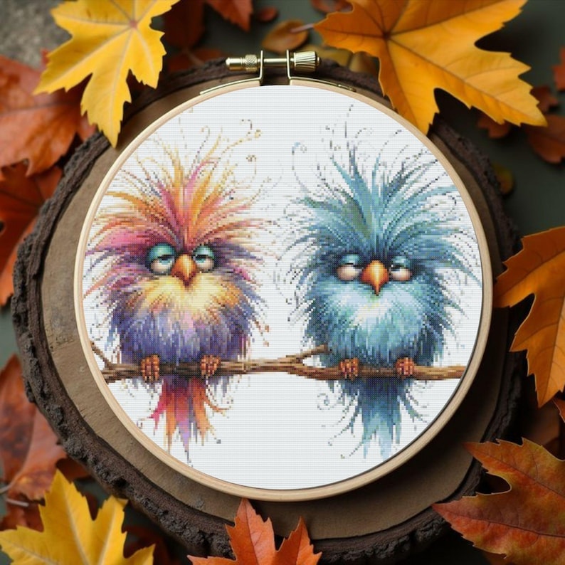 K&ouml;nnte beinhalten: Ein Kreuzstich-Stickrahmen mit zwei skurrilen V&ouml;geln auf einem Ast. Die V&ouml;gel haben farbenfrohes, flauschiges Gefieder in Lila, Orange, Gelb und Blau. Der Hintergrund umfasst Herbstbl&auml;tter.