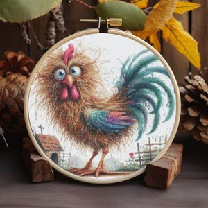 Puede incluir: Un bordado de punto de cruz de un gallo caprichoso con un cuerpo marrón esponjoso, plumas de cola azules y verdes y una cresta rosa. La obra de arte está enmarcada en un aro de bordado de madera, con una pequeña casa y una valla en el fondo.
