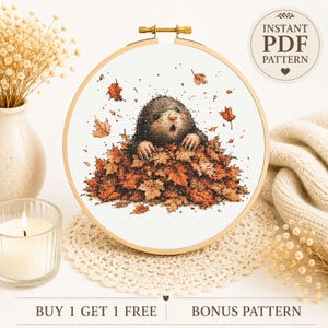 May include: A cross-stitch pattern in a wooden embroidery hoop featuring a mole emerging from a pile of autumn leaves. The design includes scattered orange and brown foliage. Text in the image reads INSTANT PDF PATTERN and BUY 1 GET 1 FREE.