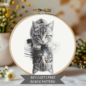 May include: Cross-stitch embroidery of a gray tabby kitten with amber eyes, reflected in water. The kitten is centered in a wooden embroidery hoop. The text "BUY 1 GET 1 FREE BONUS PATTERN" is displayed below the hoop.