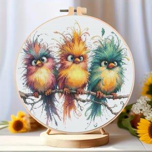 K&ouml;nnte beinhalten: Ein Kreuzstich-Stickrahmen mit drei bunten V&ouml;geln auf einem Ast. Die V&ouml;gel haben leuchtende Federn in Rosa, Orange und Gr&uuml;n. Die Stickerei ist in einem Holzrahmen gefasst und auf einem Holzst&auml;nder ausgestellt.