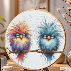K&ouml;nnte beinhalten: Ein Kreuzstich-Stickrahmen mit zwei skurrilen V&ouml;geln auf einem Ast. Die V&ouml;gel haben farbenfrohes, stacheliges Gefieder in Orange-, Rosa-, Lila- und Blaut&ouml;nen. Die Stickerei befindet sich in einem Holzrahmen.