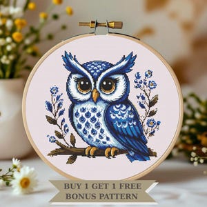 May include: Cross-stitch embroidery of a blue and white owl perched on a branch, framed in a wooden hoop. The owl has large brown eyes and is surrounded by blue floral accents. The text "BUY 1 GET 1 FREE BONUS PATTERN" is at the bottom.