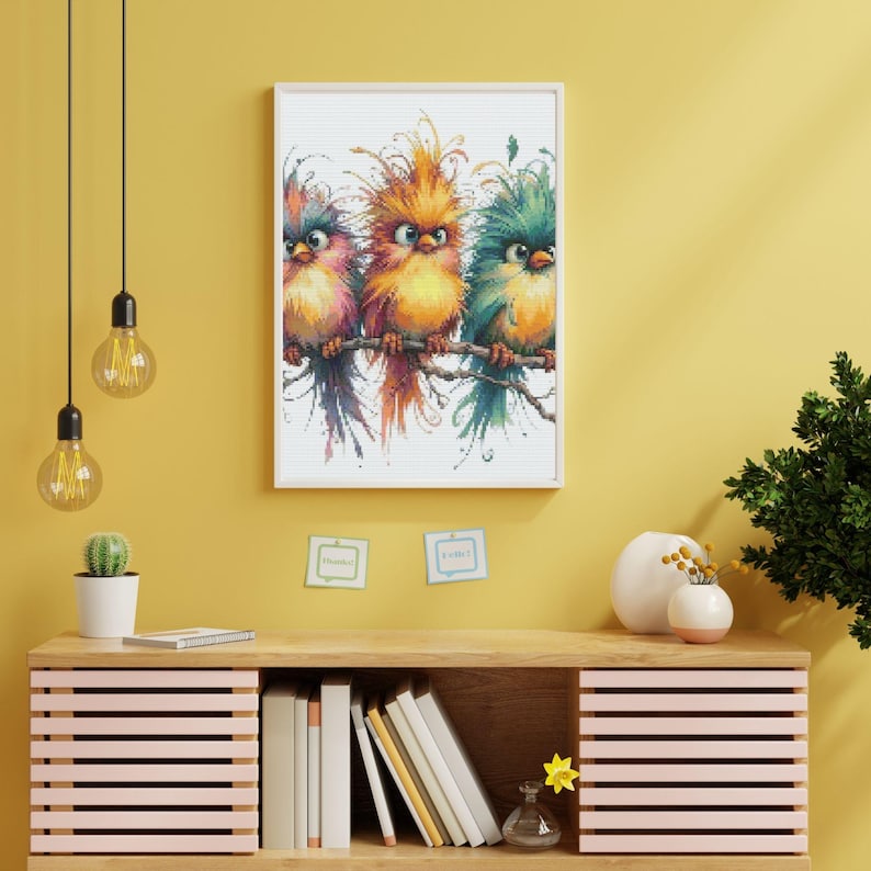 K&ouml;nnte beinhalten: Ein gerahmter Druck mit drei bunten V&ouml;geln, die auf einem Ast sitzen. Die V&ouml;gel haben ein lebendiges Gefieder in Rosa-, Orange- und T&uuml;rkist&ouml;nen. Das Kunstwerk wird an einer gelben Wand &uuml;ber einem Holzschrank mit B&uuml;chern und Dekoration ausgestellt.