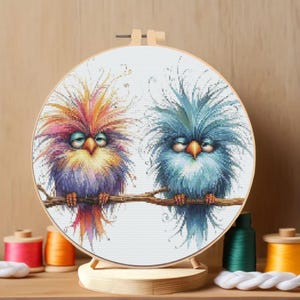 K&ouml;nnte beinhalten: Ein Kreuzstich-Stickrahmen mit zwei skurrilen V&ouml;geln auf einem Ast. Die V&ouml;gel haben ein farbenfrohes, flauschiges Gefieder in Rosa, Orange, Blau und Lila. Die Stickerei befindet sich in einem Holzrahmen, mit Garnrollen im Hintergrund.