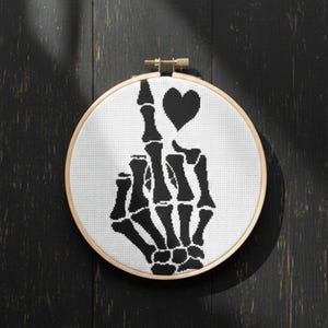 May include: A black and white cross-stitch embroidery hoop featuring a skeleton hand giving the middle finger, with a black heart above the hand. The design is on white fabric, set in a wooden hoop.