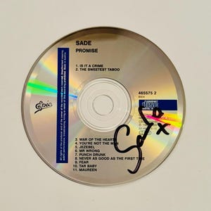 以下が含まれることがあります： Sadeの「Promise」というタイトルのCD。銀色のディスクには、「Is It a Crime」や「The Sweetest Taboo」などの曲名が黒字で記載されています。ディスクには黒いマーカーでサインが書かれています。