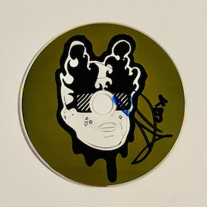 May include: A compact disc with a unique design. The disc features a black and white illustration of a face wearing sunglasses, with a green background. The design includes a signature and small star shapes.